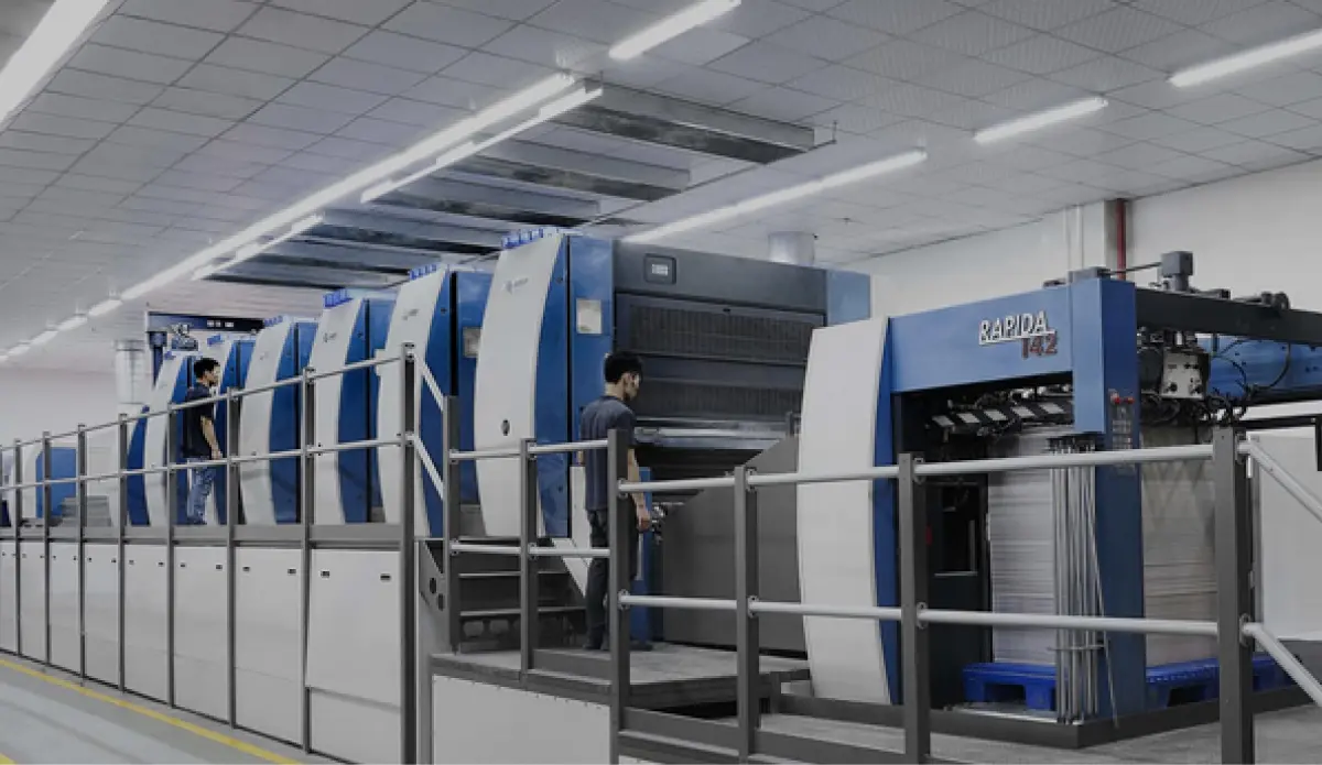 Workers operating large blue printing machinery for package manufacturing in an industrial facility.