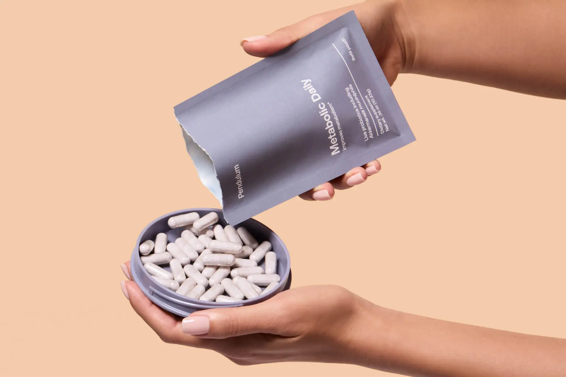 Hands pouring probiotic capsules from a Pendulum Metabolic Daily refill pouch into a reusable container.