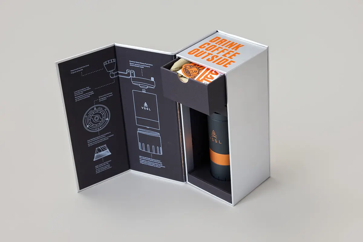Open VSSL outdoor coffee gear packaging box displaying technical diagrams on the inner lid, a top drawer with stickers reading "Drink Coffee Outside," and a cylindrical coffee grinder inside.