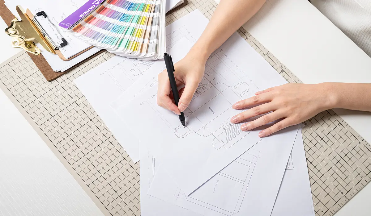 A designer drawing a structural dieline for package design next to a Pantone color guide, representing a One-stop Packaging Partner